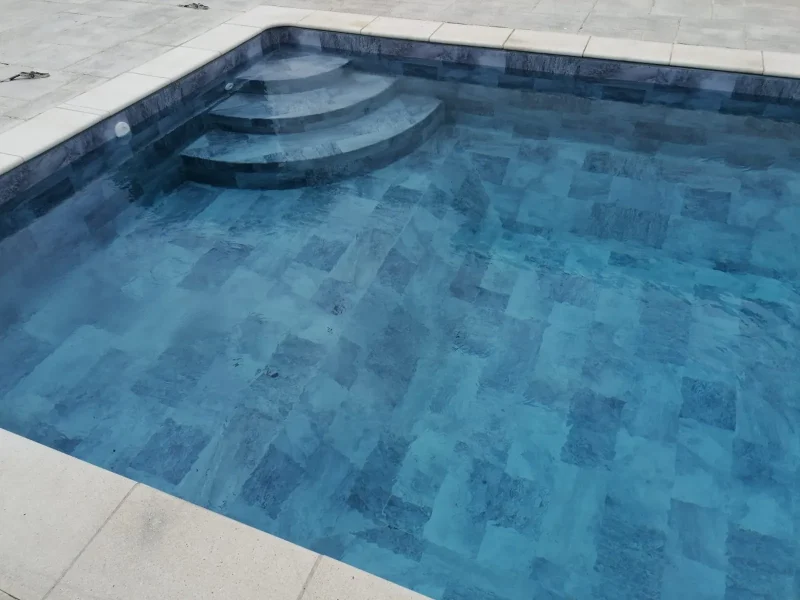 Piscine rectangulaire remplie d'eau claire avec un revêtement en membrane armée aspect pierre grise, un escalier arrondi d'angle et des margelles beiges